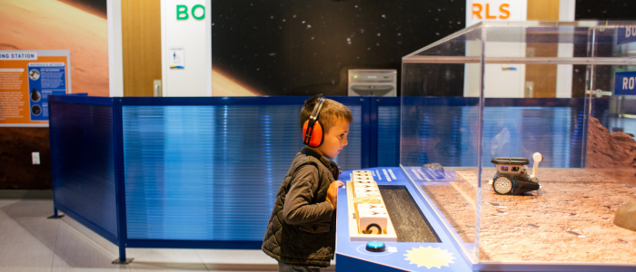 A child wearing headphones observes a robotic rover inside a glass display in a space-themed exhibit.