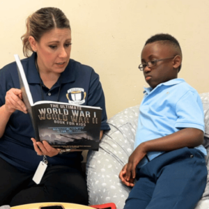 A woman reads a book titled "The Ultimate World War II Book for Kids" to a young boy seated on a bed, engaged and attentive.