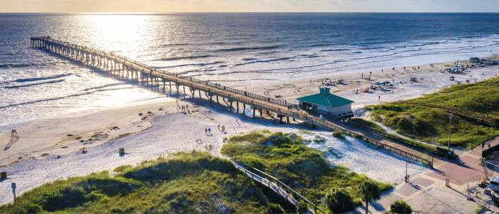 Aerial view of a long pier leading to the ocean, with sunlit waves, sandy beach, and people enjoying the seaside.