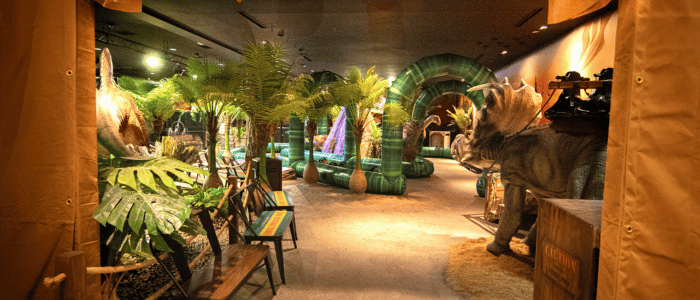 A vibrant exhibits space with lush greenery, dinosaur statues, and colorful lighting, creating a prehistoric-themed atmosphere.