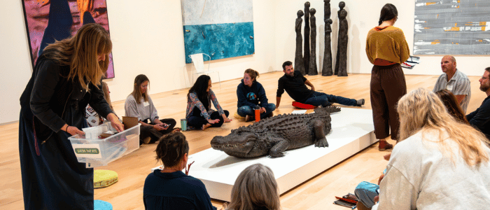 The Dennos Museum A group of people sit on the floor in an art gallery, observing a large crocodile displayed on a pedestal amidst various artworks.