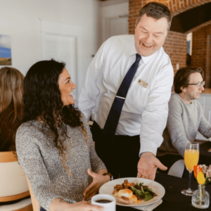 Server presenting a plated meal to a seated woman in a cozy restaurant setting with other diners in the background.