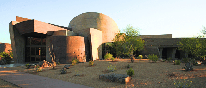 Modern, dome-shaped building with curved concrete walls surrounded by desert landscaping and a clear blue sky.