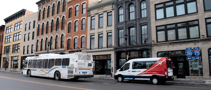 City street with historic brick buildings, a public transit bus, a transit shuttle van, and highway direction signs.