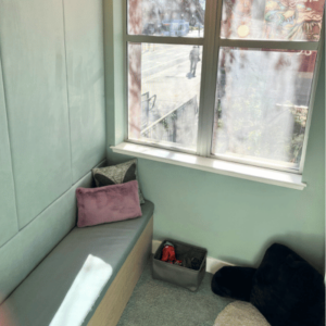 Cozy corner with a built-in bench, pink and patterned cushions, a window overlooking a street, and a basket on a carpeted floor.