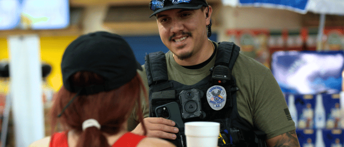 Police officer wearing a body camera and uniform talks to a woman with red hair and a black cap in an indoor setting.