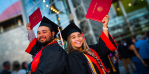 Two cheerful members of the Benedictine University graduating class dressed in cap and gown with diploma books in hand