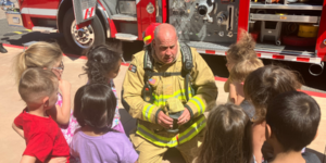 Firefighter surrounded by a group of children outside with a fire engine in the background.