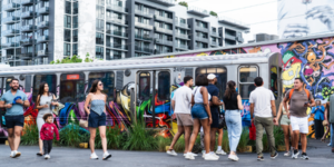 wynwood_walls_v2 Crowd of people at an outdoor exhibit at Wynwood walls with graffitied walls and subway car