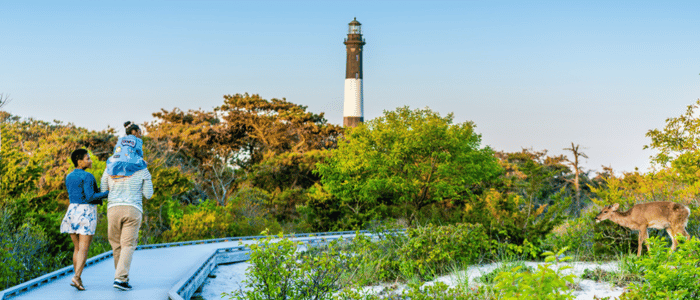 Family walking on a boardwalk near green trees and a lighthouse, with a deer standing on the right side in a natural setting.