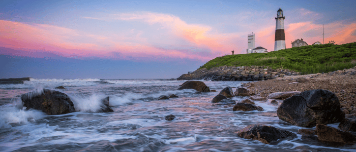 Seaside view of a lighthouse on a rocky shore at sunset with waves crashing and a colorful sky.