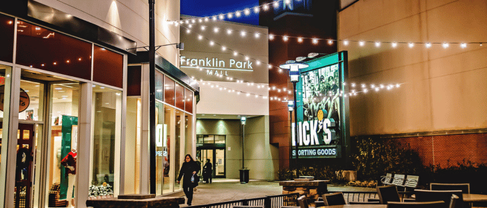 Outdoor nighttime view of Franklin Park Mall entrance with string lights, shoppers, benches, and a lit store sign.