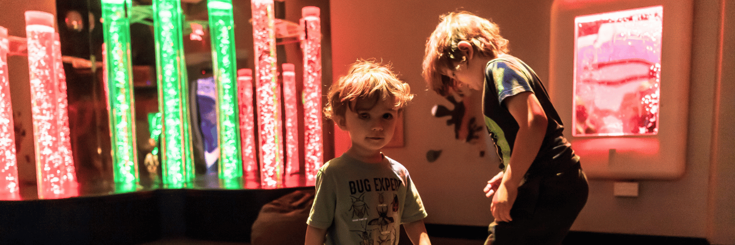 Two children explore an interactive light exhibit in a dimly lit children’s museum.