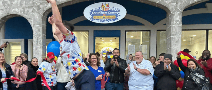 Crowd applauding outside Vincent’s Clubhouse Enrichment Center during a ribbon-cutting event with a person jumping in front.