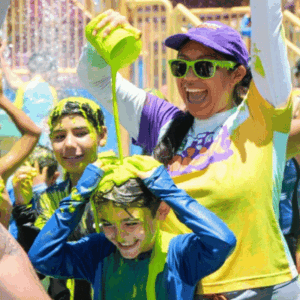 Woman in sunglasses pours water from a green cup over a smiling child at a colorful outdoor water festival.