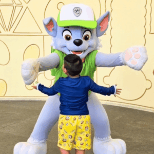 Child in a blue shirt and yellow shorts hugs a large blue-and-white cartoon wolf mascot wearing a green vest and white cap.
