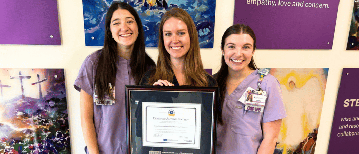 Three smiling women in purple scrubs holding a framed Certified Autism Center® award in a colorful art-decorated room.
