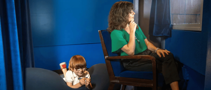A woman in a green top sitting on a chair looking out a window, while a boy with glasses lies on a beanbag nearby.