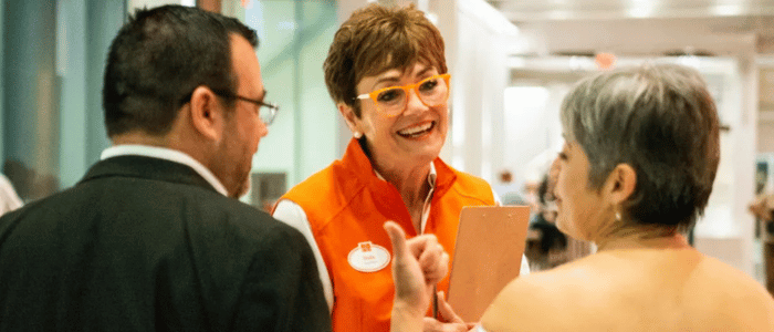 Three people are engaged in a cheerful conversation in a bright indoor setting, with one woman wearing an orange vest and glasses.