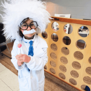 Child dressed as Albert Einstein with wild white wig, glasses, and lab coat, holding a lollipop next to a large Connect Four game.