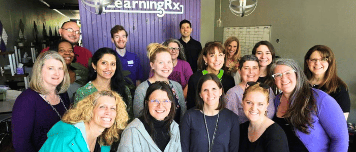 LearningRx Chattanooga (2) Group photo of 18 smiling adults standing indoors in front of a purple wall with "LearningRx" sign.