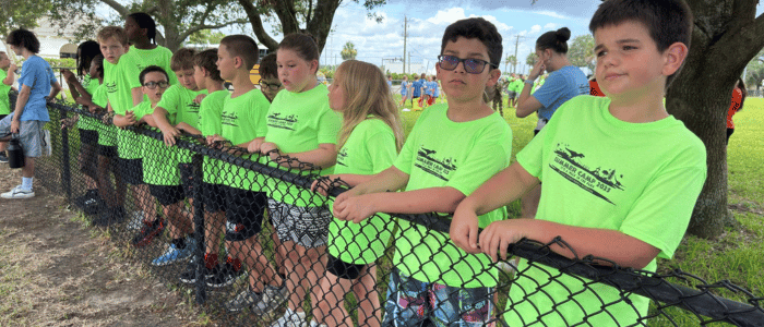 St. Johns County Parks and Recreation (1) A group of children in bright green T-shirts stand along a black chain-link fence outdoors under trees on a sunny day.