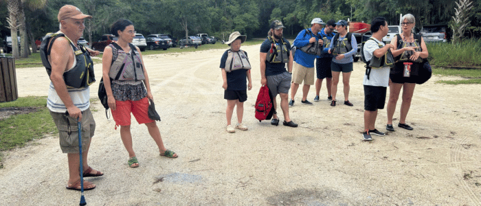 St. Johns County Parks and Recreation (2) A group of people wearing life jackets stand on a gravel path near parked cars, preparing for an outdoor water activity.