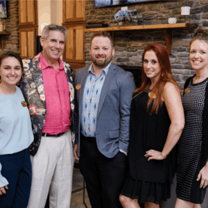 Five adults pose together indoors in front of a stone fireplace and wooden cabinetry, smiling at the camera.