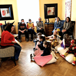 Children sit on the floor listening to a woman read aloud, while adults watch in a cozy room.